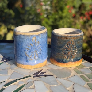 May include: Two cylindrical ceramic cups with a glazed finish. The left cup is light blue with a leaf-like embossed pattern, while the right cup is a darker blue with a diamond-shaped embossed pattern. Both have unglazed tan bases.