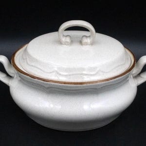 May include: A round, off-white ceramic casserole dish with a speckled finish and a matching lid. The dish features two side handles and a central loop handle on the lid. A thin, brown decorative band encircles the rim of the base and the lid.