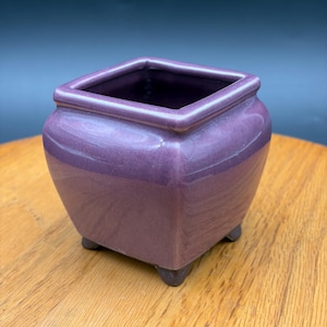 May include: A square-shaped ceramic planter with a glossy purple glaze. The vessel features a wide, open top with a thick rim, a slightly tapered body, and four small, dark-colored feet at the base that elevate the container slightly.