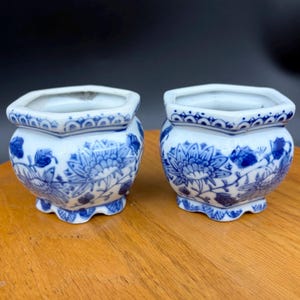 May include: Two small, matching ceramic pots with a blue and white floral design. The pots have a hexagonal shape with a scalloped rim and a detailed flower pattern. The pots are placed on a wooden surface.