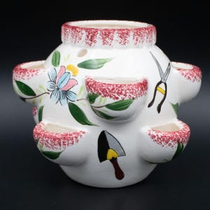 May include: White ceramic strawberry pot with multiple planting pockets. The pot features hand-painted floral and gardening tool designs, including a trowel and shears. The rim and pocket edges have a red speckled pattern.