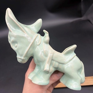 May include: A light mint green ceramic donkey figurine featuring a glossy finish, a molded saddle, and a bridle. The donkey is depicted in a standing position with its head tilted downward. A wooden ruler is partially visible at the bottom right.