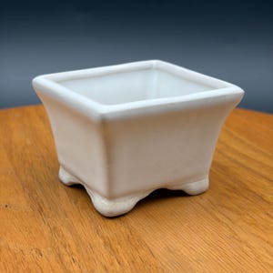 May include: A square white ceramic planter with a smooth finish and a flared rim. The container features four small, rounded feet at the base, providing a stable foundation for this open-top vessel designed for holding small plants.
