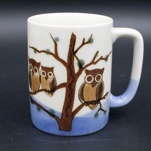 May include: White ceramic mug featuring a brown tree with owls perched on its branches. The mug has a blue base and a white handle. The owls are brown and tan with large, round eyes. A decorative item for the kitchen.