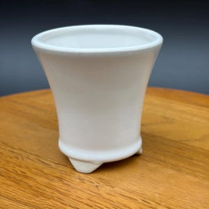 May include: A small, off-white ceramic planter with a smooth, matte finish. The vessel features a flared rim, a tapered body, and stands on three short, integrated feet. The interior is hollow and the exterior surface is plain and unadorned.