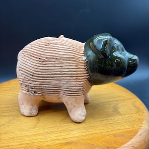 May include: A ceramic pig figurine featuring a textured, unglazed terracotta body with horizontal ridges and a smooth, glossy dark green glazed head. The animal figure stands on four short legs and has a rounded snout and small ears.