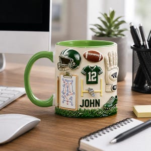 Puede incluir: Una taza de cerámica color crema con asa y borde verdes, decorada con adornos temáticos de fútbol americano. Presenta un casco verde, un balón de fútbol, una camiseta con el número 12 y el nombre "JOHN".