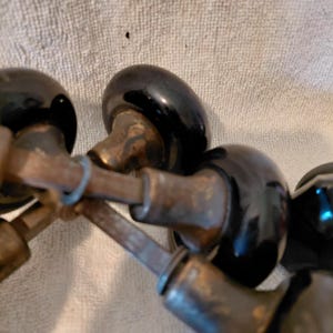 May include: A collection of vintage door knobs featuring smooth, round, black glass or ceramic heads attached to tarnished, weathered brass-toned metal shanks and square-shaped spindles. The hardware components show visible signs of age and oxidation.