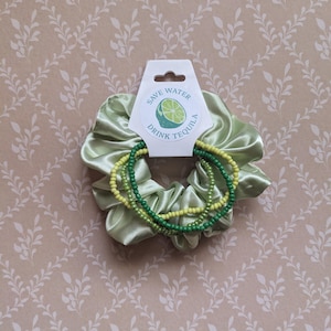 May include: A light green satin scrunchie with three beaded bracelets in shades of green and yellow. The scrunchie has a tag that reads "Save Water Drink Tequila" with a lime graphic. The background is a light brown with a leaf pattern.