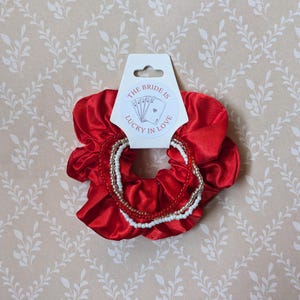 May include: A bright red satin scrunchie paired with three beaded bracelets featuring white, red, and metallic gold beads. A white display card is attached, printed with the text THE BRIDE IS LUCKY IN LOVE and an illustration of playing cards.