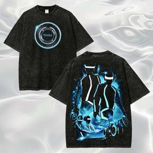 May include: A black acid-wash short-sleeve t-shirt. The front features a circular blue glowing Tron logo. The back displays a large graphic of figures in glowing blue suits and helmets with light cycles, set against a dark, energetic background.