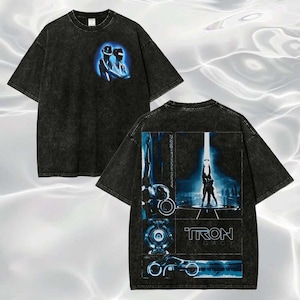 May include: A black acid-wash t-shirt featuring a small graphic of two figures on the front chest. The back displays a large collage of Tron Legacy imagery, including characters, a light cycle, and the text Tron Legacy in a blue and black color scheme.