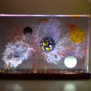 May include: A rectangular resin block light featuring a space-themed design with swirling white nebula clouds, glitter, and five distinct circular planet-like spheres in black, blue, yellow, and white, mounted on a dark wooden base.