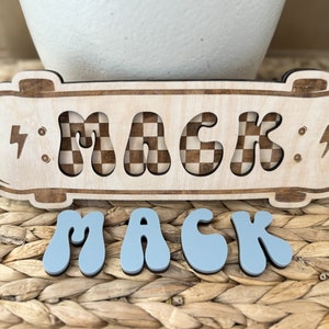 May include: A wooden skateboard-shaped sign with the name "MACK" spelled out in a checkered pattern. The sign is sitting on a woven wicker surface. Below the sign are four blue letters that spell out "MACK" in a wavy font.
