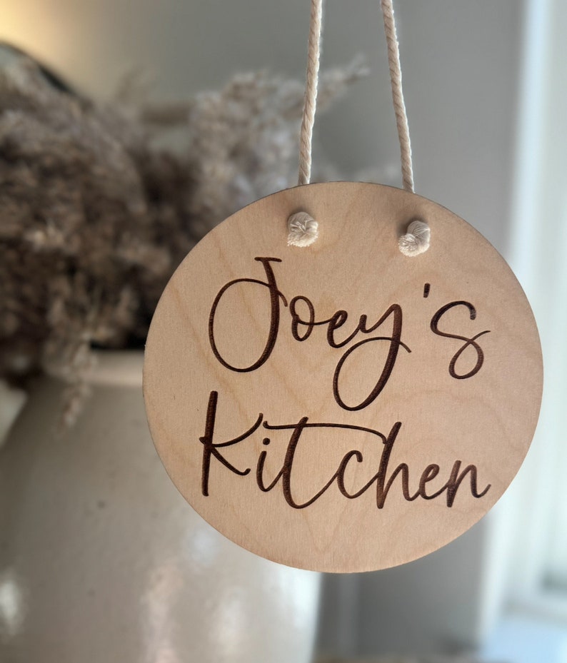 K&ouml;nnte beinhalten: Ein kreisf&ouml;rmiges Schild aus hellem Holz mit dem eingravierten Text "Joey's Kitchen" in einer dunklen Schreibschrift. Das Schild ist an einem d&uuml;nnen wei&szlig;en Seil aufgeh&auml;ngt, das durch zwei L&ouml;cher an der Oberseite gezogen wurde.