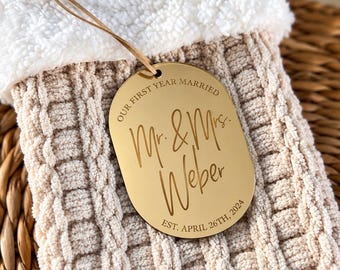May include: A gold-colored oval acrylic ornament with a tan leather cord, featuring the engraved text "OUR FIRST YEAR MARRIED", "Mr. & Mrs. Weber", and "EST. APRIL 26TH, 2024", resting on a beige cable-knit stocking with a white faux-fur cuff.