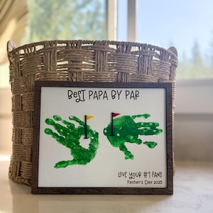 May include: A framed handprint art piece with the text "Best Papa By Par" and "Love Your #1 Fans Father's Day 2023". The handprints are green and represent golf putting greens with flags.