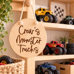 K&ouml;nnte beinhalten: Ein rundes, helles Holzschild, das an einem wei&szlig;en Seil h&auml;ngt und den dunkelbraun gravierten Text "Crew's Monster Trucks" in Schreibschrift zeigt. Das Schild ist kreisf&ouml;rmig und hat zwei kleine Knoten, die das Seil oben sichern.
