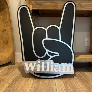 May include: Black wooden hand sign with white outline in the shape of a rock and roll hand gesture. The name "William" is written in white letters on a silver background.