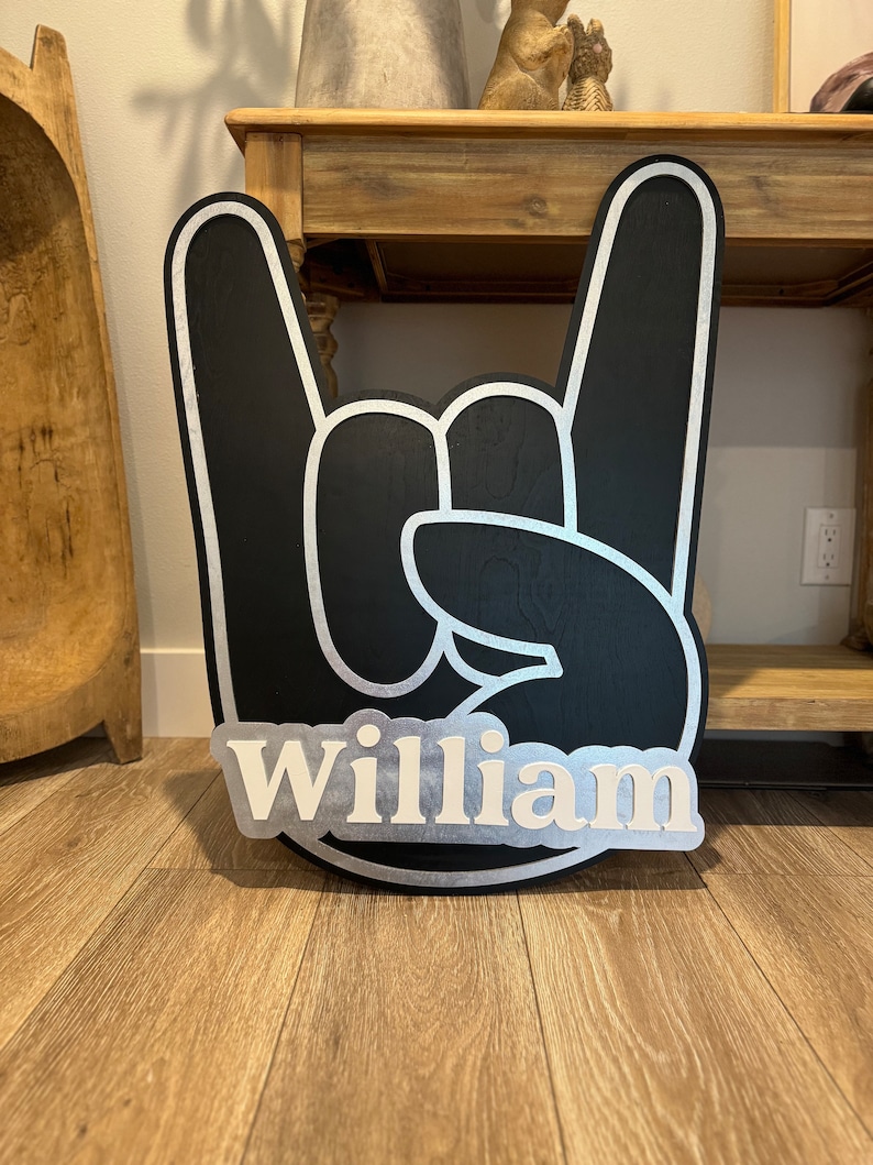 May include: Black wooden hand sign with white outline in the shape of a rock and roll hand gesture. The name "William" is written in white letters on a silver background.