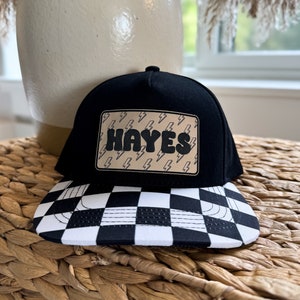 May include: Black and white checkered baseball cap with a black crown and a brown patch with the name "HAYES" in white letters and a lightning bolt pattern.