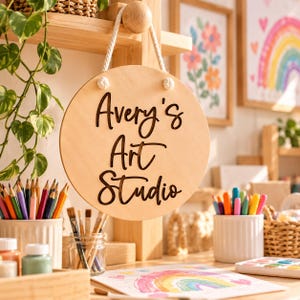 K&ouml;nnte beinhalten: Ein rundes, helles Holzschild, das an einem wei&szlig;en Seil h&auml;ngt und den schwarzen, lasergeschnittenen Schriftzug Avery's Art Studio tr&auml;gt. Das Schild befindet sich in einem Kreativbereich mit Buntstiften, Pinseln und einer Regenbogenzeichnung.