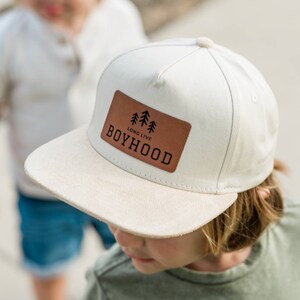 May include: A white and brown baseball cap with a brown leather patch that says "Long Live Boyhood" with a small graphic of three trees.