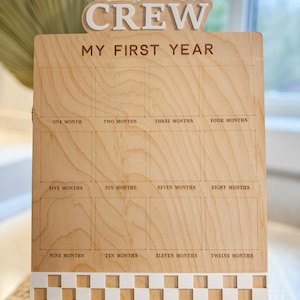 May include: A wooden sign with the words "CREW MY FIRST YEAR" and a grid of 12 squares, each labeled with a month of the year. The sign is decorated with a checkered border.