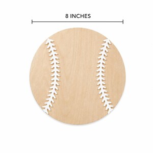 May include: A circular wooden cutout shaped like a baseball, featuring a light tan natural wood grain finish and two white curved stitched patterns on the surface. The item measures 8 inches in diameter as indicated by the text and line above.