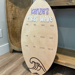 May include: A wooden surfboard-shaped sign with the text "kenzie's FIRST WAVE" in purple and white lettering. The sign has 12 blank squares labeled with numbers one through twelve.