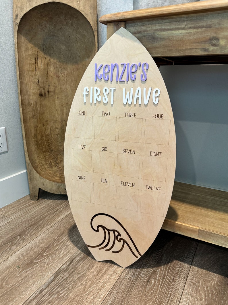 May include: A wooden surfboard-shaped sign with the text "kenzie's FIRST WAVE" in purple and white lettering. The sign has 12 blank squares labeled with numbers one through twelve.