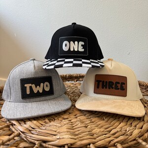 May include: Three baseball caps with numbers one, two, and three on them. The caps are black, gray, and beige. The numbers are in white, black, and brown.