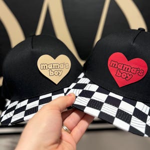 May include: Two black baseball caps with a checkered pattern on the brim. One cap has a red heart with the text "mama's boy" on it. The other cap has a tan heart with the same text.