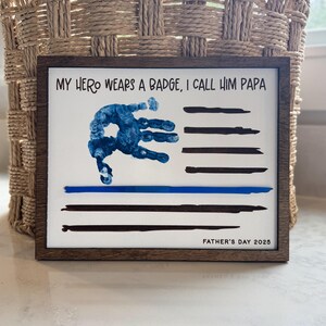 May include: A rectangular wooden framed sign with a white background featuring the text MY HERO WEARS A BADGE, I CALL HIM PAPA at the top and FATHER'S DAY 2025 at the bottom. A blue handprint and black horizontal stripes form a flag design.