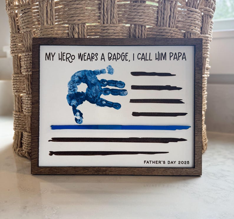 May include: A rectangular wooden framed sign with a white background featuring the text MY HERO WEARS A BADGE, I CALL HIM PAPA at the top and FATHER'S DAY 2025 at the bottom. A blue handprint and black horizontal stripes form a flag design.