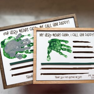 May include: Two rectangular wooden framed signs with white backgrounds. The text reads "OUR HERO WEARS CAMO, WE CALL HIM DADDY!" and "MY HERO WEARS CAMO, I CALL HIM DADDY!" featuring green handprints, dark brown painted stripes, and "THANK YOU FOR KEEPING ME SAFE!"
