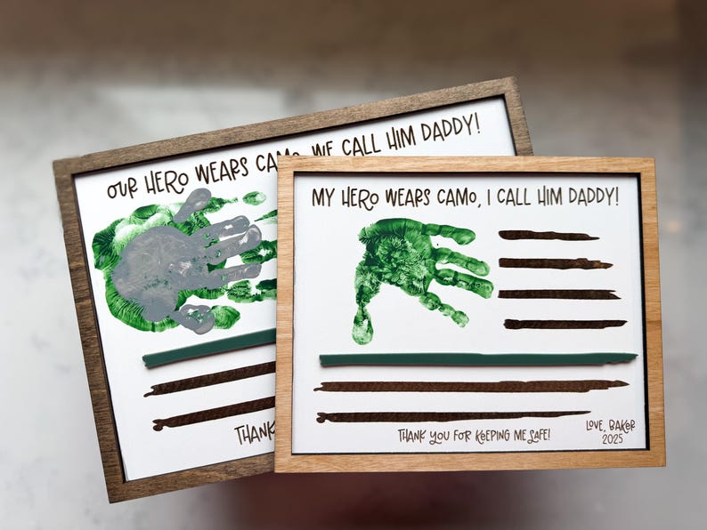 May include: Two rectangular wooden framed signs with white backgrounds. The text reads "OUR HERO WEARS CAMO, WE CALL HIM DADDY!" and "MY HERO WEARS CAMO, I CALL HIM DADDY!" featuring green handprints, dark brown painted stripes, and "THANK YOU FOR KEEPING ME SAFE!"