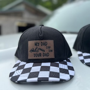 May include: Black and white checkered baseball cap with a brown patch that says "My Dad Can Tow Your Dad" and a small image of a tow truck and car.