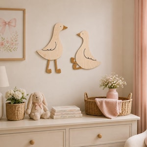 May include: Two light-colored wooden wall decorations shaped like geese, one standing and one sitting, mounted on a wall. The standing goose has long legs and the sitting goose has bent legs, both featuring simple black line details for wings and eyes.
