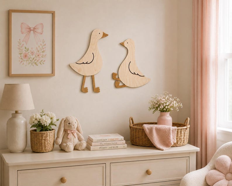 May include: Two light-colored wooden wall decorations shaped like geese, one standing and one sitting, mounted on a wall. The standing goose has long legs and the sitting goose has bent legs, both featuring simple black line details for wings and eyes.