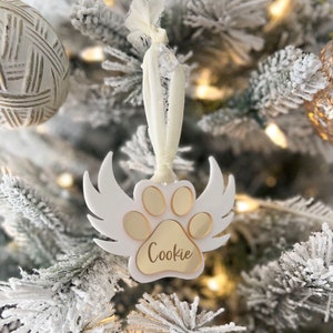 May include: White ceramic Christmas ornament shaped like a paw print with angel wings. The paw print has gold accents and the name "Cookie" is inscribed on the bottom.