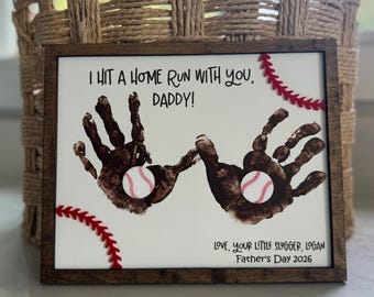 Puede incluir: Un regalo enmarcado para el Día del Padre con arte de huellas de manos. La obra presenta dos huellas de manos marrones con pelotas de béisbol, costuras rojas y el texto "I HIT A HOME RUN WITH YOU, DADDY!" y "LOVE, YOUR LITTLE SLUGGER, LOGAN Father's Day 2026."