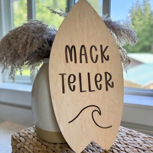 Könnte beinhalten: Holzschild in Form eines Surfbretts mit dem Namen "Mack Teller" und einem Wellendesign. Das Schild ist hellbraun und hat eine natürliche Holzmaserung.