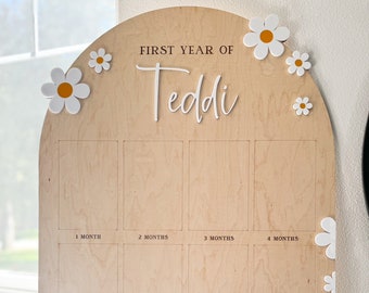 May include: A wooden arch-shaped growth chart with the text "FIRST YEAR OF Teddi" and 12 blank spaces for photos. The chart is decorated with white daisies with yellow centers.