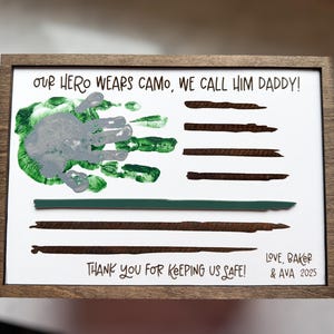 May include: A rectangular wooden frame containing a white sign with the text "OUR HERO WEARS CAMO, WE CALL HIM DADDY!" and "THANK YOU FOR KEEPING US SAFE!". It features a grey and green handprint, brown painted stripes, and "LOVE, BAKER & AVA 2025".