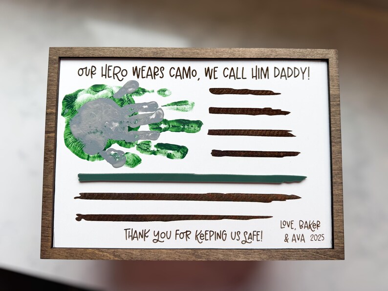 May include: A rectangular wooden frame containing a white sign with the text "OUR HERO WEARS CAMO, WE CALL HIM DADDY!" and "THANK YOU FOR KEEPING US SAFE!". It features a grey and green handprint, brown painted stripes, and "LOVE, BAKER & AVA 2025".