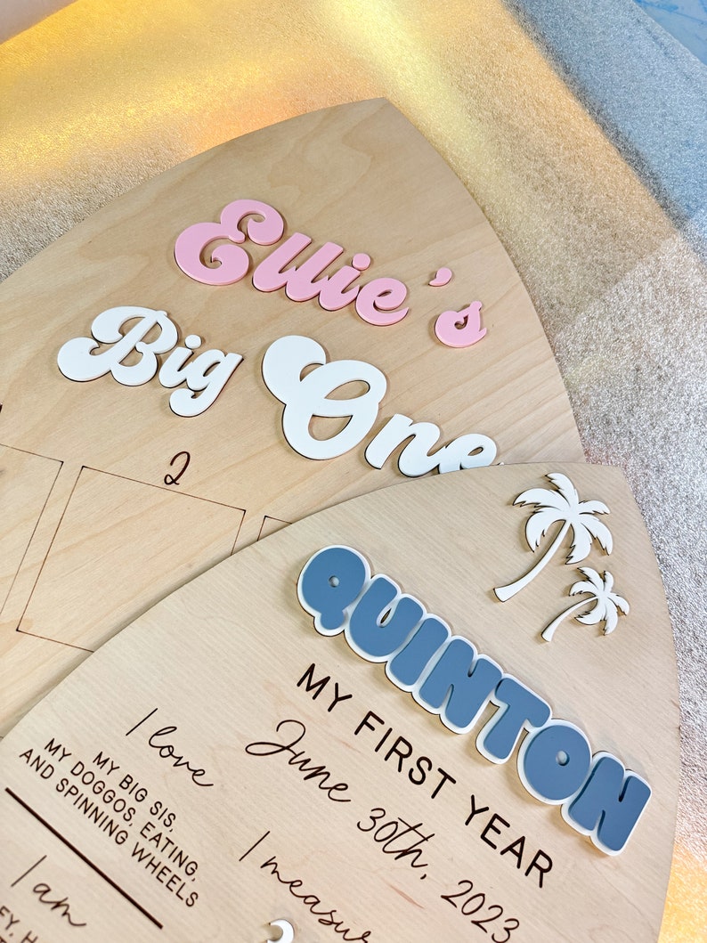 May include: A wooden sign with the text "Ellie's Big One" in pink and white letters. The sign also includes the text "My First Year June 30th, 2023" and a blue and white palm tree.