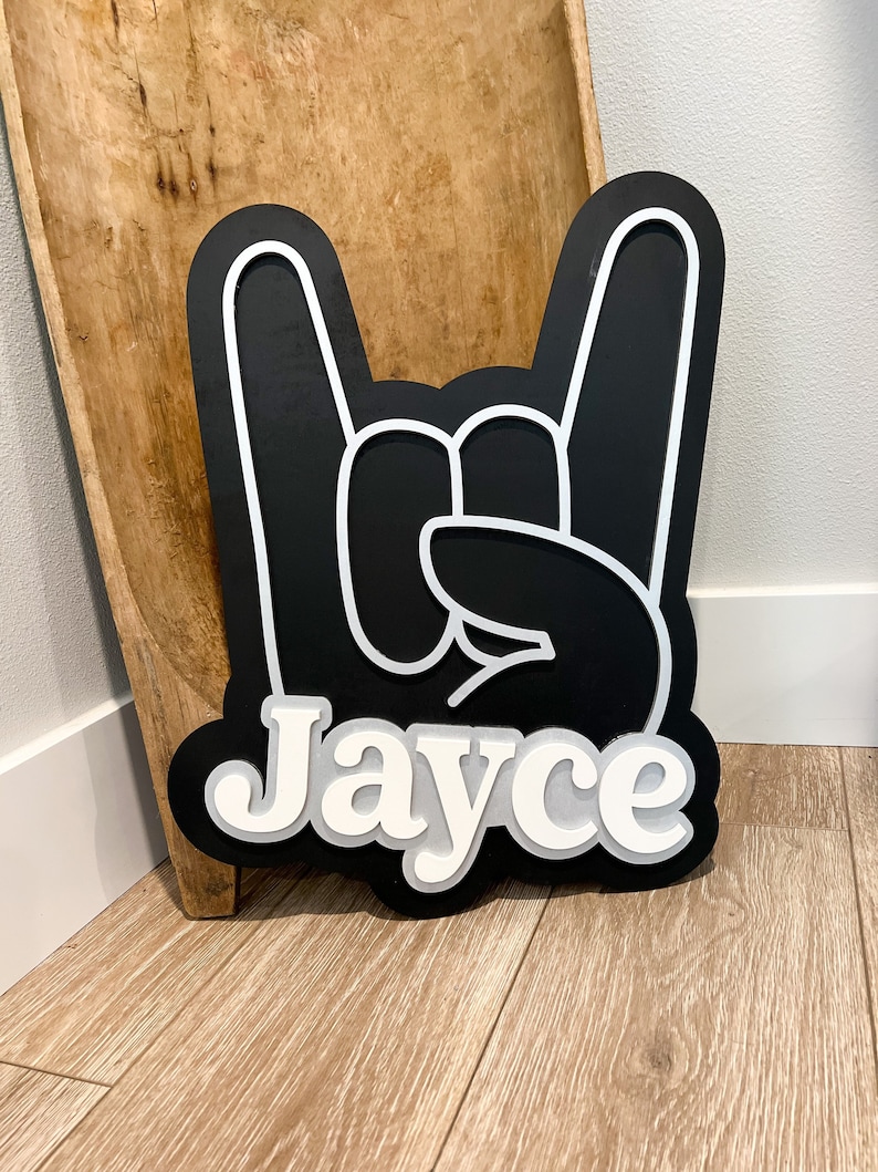 May include: Black wooden sign with a white outline of a hand making the rock and roll hand gesture. The name "Jayce" is written in white letters below the hand.