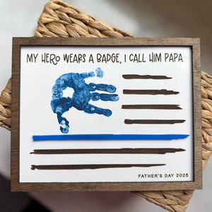 May include: A rectangular wooden frame containing a white print with the text MY HERO WEARS A BADGE, I CALL HIM PAPA and FATHER'S DAY 2025. The design features a blue handprint and horizontal black lines with a single thin blue line.
