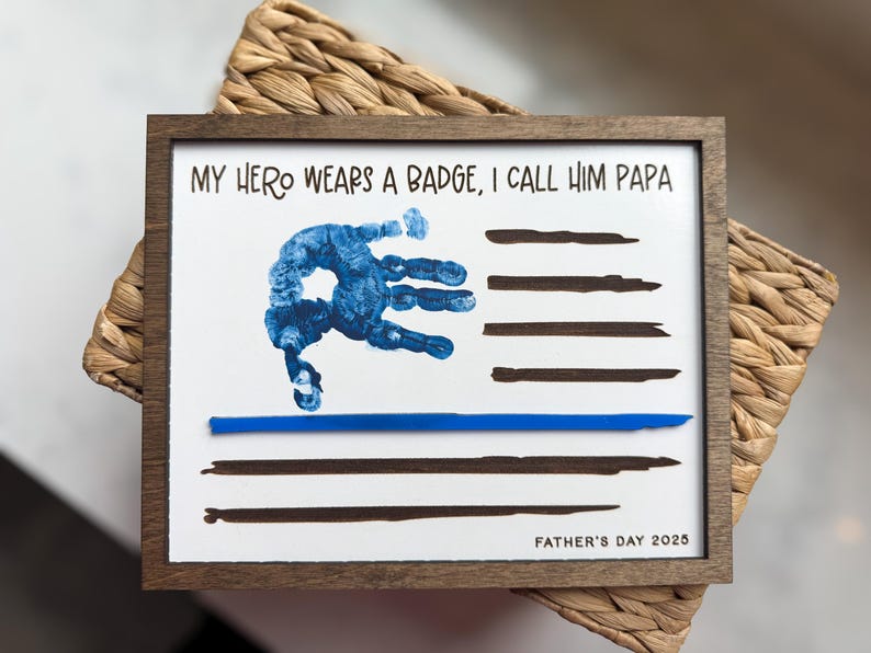 May include: A rectangular wooden frame containing a white print with the text MY HERO WEARS A BADGE, I CALL HIM PAPA and FATHER'S DAY 2025. The design features a blue handprint and horizontal black lines with a single thin blue line.